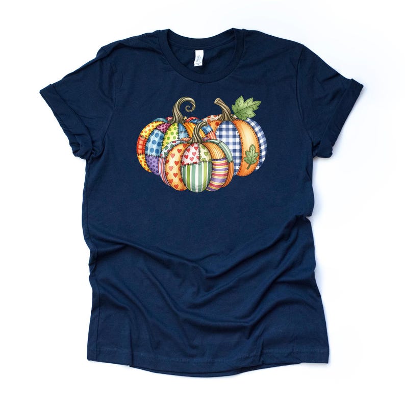 Pu&ograve; includere: Una maglietta blu navy con una grafica di tre zucche. Le zucche hanno un design patchwork con vari motivi e colori, tra cui pois, cuori e strisce. Le zucche sono disposte in fila, con foglie verdi e steli ricci.