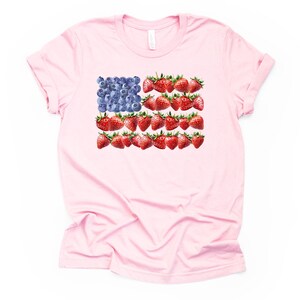 May include: A light pink t-shirt featuring a design of the American flag made of blueberries and strawberries. The blueberries form the blue field, and the strawberries create the red stripes. The shirt has short sleeves and a crew neck.
