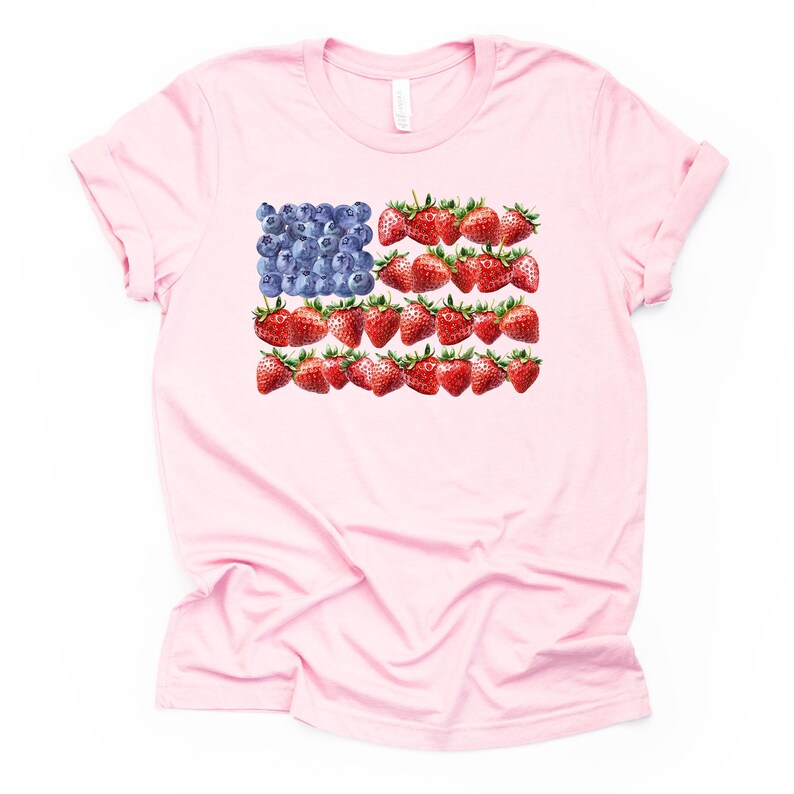 May include: A light pink t-shirt featuring a design of the American flag made of blueberries and strawberries. The blueberries form the blue field, and the strawberries create the red stripes. The shirt has short sleeves and a crew neck.