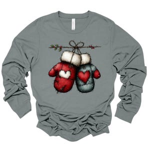 May include: A long-sleeved gray shirt featuring a graphic of two mittens tied together with a string. One mitten is red with a white heart, and the other is blue with a red heart. The mittens are decorated with holly.