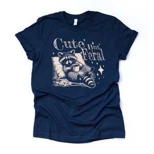 May include: A navy blue t-shirt with a graphic of a raccoon holding a mug and the text "Cute But Feral".