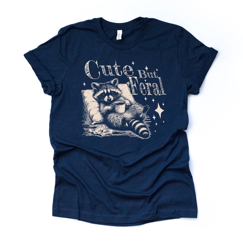 May include: A navy blue t-shirt with a graphic of a raccoon holding a mug and the text "Cute But Feral".