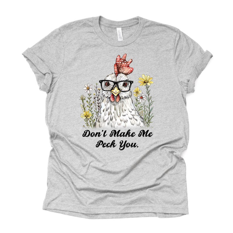 May include: A light gray t-shirt featuring a whimsical illustration of a chicken wearing glasses, surrounded by flowers. The text "Don't Make Me Peck You" is printed below the image. The shirt is made of a soft material.