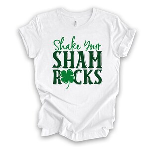 Puede incluir: Camiseta blanca con texto verde que dice "Shake Your Shamrocks" con un tr&eacute;bol de cuatro hojas verde.