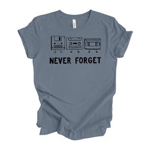 May include: A blue t-shirt featuring a black graphic of a floppy disk, cassette tape, and VHS tape with legs. The text "NEVER FORGET" is printed below the graphics in black.