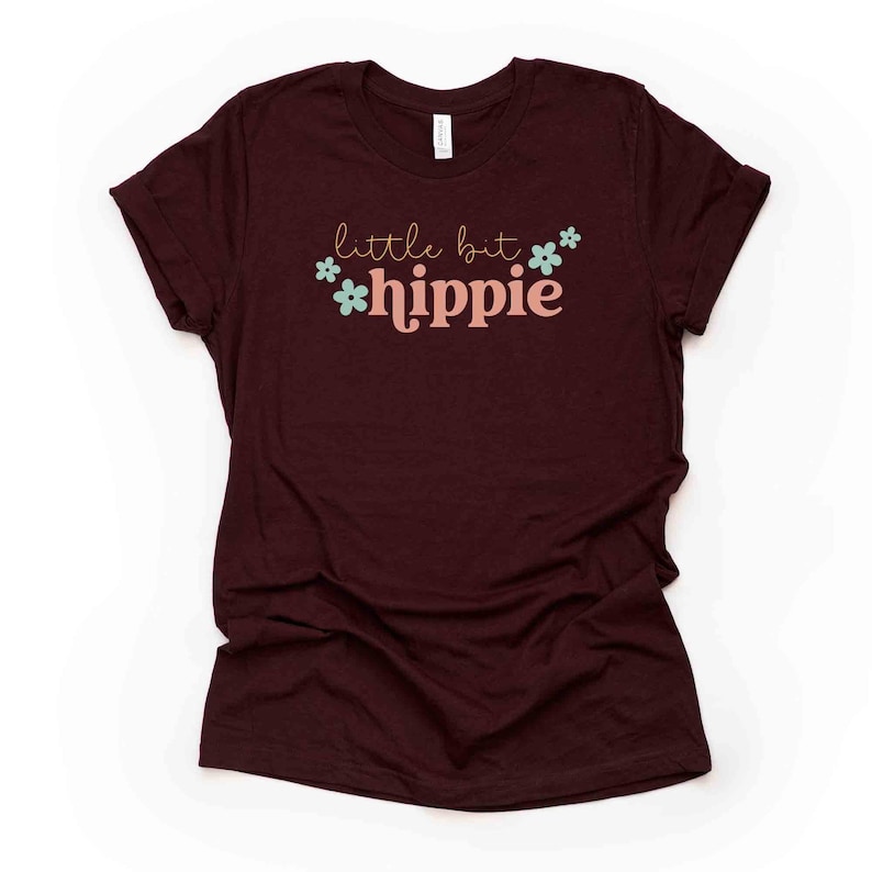 Peut inclure: Un t-shirt marron avec le texte "little bit hippie" en lettres roses et vertes avec de petits motifs de fleurs.