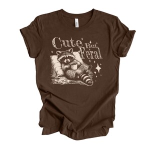 May include: Brown t-shirt with a graphic of a raccoon holding a cup of coffee and the text "Cute But Feral".