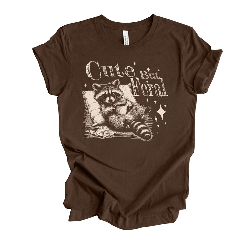 May include: Brown t-shirt with a graphic of a raccoon holding a cup of coffee and the text "Cute But Feral".