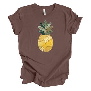 May include: A brown t-shirt featuring a patchwork pineapple design. The pineapple has yellow, green, and cream-colored fabric pieces. The shirt is made of a soft material and has short sleeves.