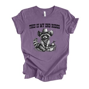Pode incluir: Camiseta roxa com uma ilustra&ccedil;&atilde;o em preto e branco de um guaxinim usando um chap&eacute;u de cowboy e montando uma sela. O texto "This is my 2nd Rodeo" est&aacute; impresso na camiseta.