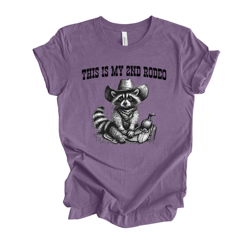 Pode incluir: Camiseta roxa com uma ilustra&ccedil;&atilde;o em preto e branco de um guaxinim usando um chap&eacute;u de cowboy e montando uma sela. O texto "This is my 2nd Rodeo" est&aacute; impresso na camiseta.