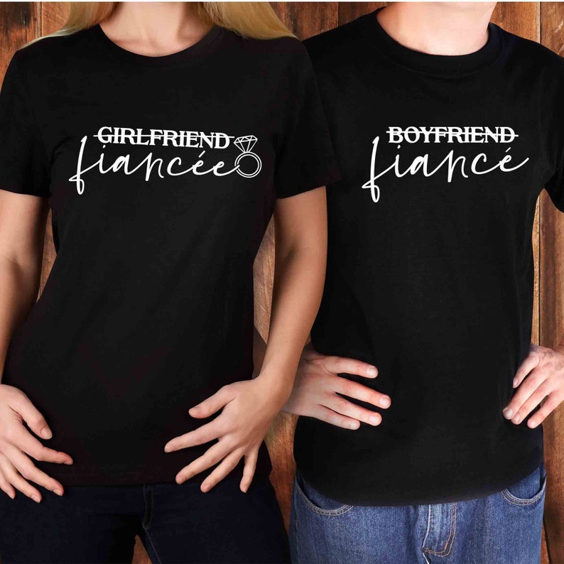 May include: Two black t-shirts with white text. The shirt on the left says "Girlfriend Fianc&eacute;e" with a diamond ring graphic. The shirt on the right says "Boyfriend Fianc&eacute;".