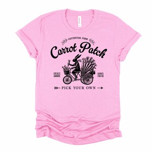 May include: Pink t-shirt with a black graphic of a rabbit riding a bicycle with a basket full of carrots. The text "Cottontail Farm Carrot Patch" is above the image, and "Locally Grown" and "Always Fresh" are on either side of the rabbit. The text "Pick Your Own" is below the image.