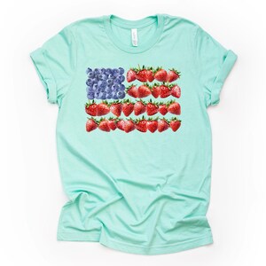 May include: A light turquoise t-shirt featuring a unique design of the American flag. The flag's stars are represented by blueberries, and the stripes are made of red strawberries. The shirt is a casual, everyday wear item.