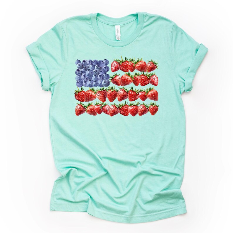 May include: A light turquoise t-shirt featuring a unique design of the American flag. The flag's stars are represented by blueberries, and the stripes are made of red strawberries. The shirt is a casual, everyday wear item.