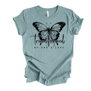 Peut inclure: Un t-shirt bleu clair avec un motif de papillon noir et le texte "transform&eacute; par l'amour de Dieu".