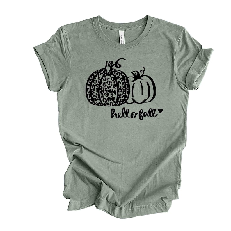 May include: Sage green t-shirt featuring a black graphic of two pumpkins. One pumpkin has a leopard print design, and the other is a solid outline. The words "hello fall" are written below the pumpkins.