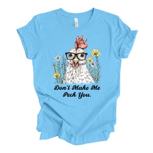 May include: Light blue t-shirt featuring a whimsical illustration of a chicken wearing glasses and surrounded by flowers. The text "Don't Make Me Peck You" is printed below the image. The shirt is made of a soft material.