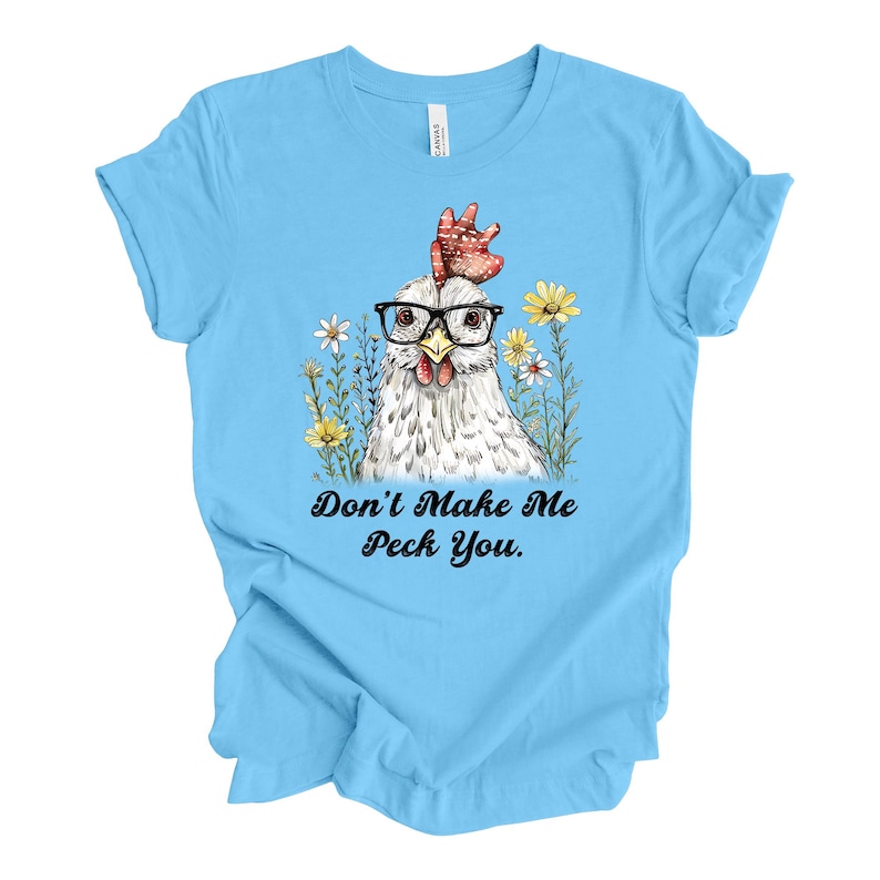 May include: Light blue t-shirt featuring a whimsical illustration of a chicken wearing glasses and surrounded by flowers. The text "Don't Make Me Peck You" is printed below the image. The shirt is made of a soft material.