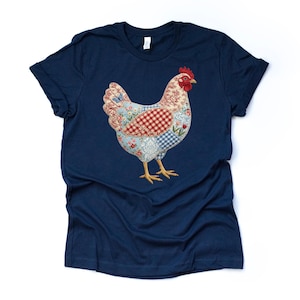 May include: Navy blue t-shirt featuring a colorful patchwork chicken design. The chicken has a red comb, and its body is made up of floral, gingham, and patterned fabric pieces. The shirt is a classic crew neck style.