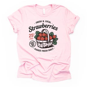 May include: A light pink t-shirt featuring a graphic of a basket filled with strawberries. The text reads "FRESH & LOCAL Strawberries" with "25¢ /PINT" and "PICKED FRESH DAILY". The design includes green leaves and a "SWEET JUICY RIPE" badge.
