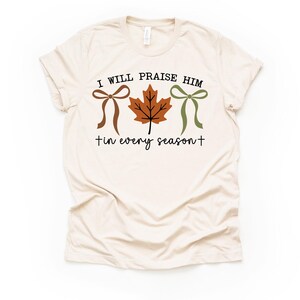 May include: Cream-colored t-shirt with the text "I WILL PRAISE HIM in every season" in black script. The design includes a brown bow, a brown leaf, and a green bow.
