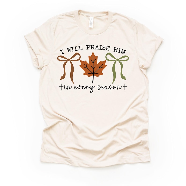 May include: Cream-colored t-shirt with the text "I WILL PRAISE HIM in every season" in black script. The design includes a brown bow, a brown leaf, and a green bow.