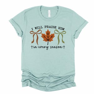 May include: Light blue t-shirt with the text "I WILL PRAISE HIM tin every season+". The shirt features a brown bow, a brown and orange leaf, and a green bow.