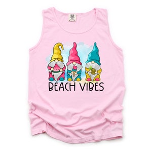 Pu&ograve; includere: Canotta rosa senza maniche con una grafica di tre gnomi che tengono dolcetti estivi. Gli gnomi indossano cappelli colorati e sono in piedi davanti a uno scenario di spiaggia. Il testo "Beach Vibes" &egrave; stampato sotto gli gnomi.