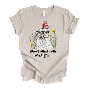 May include: A light beige t-shirt featuring a whimsical illustration of a chicken wearing glasses and surrounded by flowers. The text "Don't Make Me Peck You" is printed below the image in a playful font. The shirt is made of soft material.
