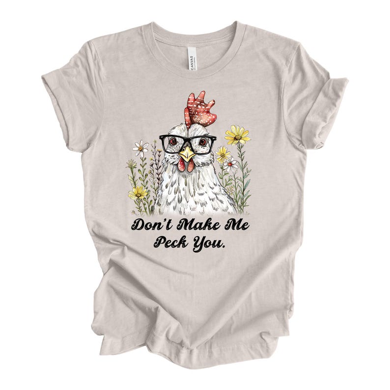 May include: A light beige t-shirt featuring a whimsical illustration of a chicken wearing glasses and surrounded by flowers. The text "Don't Make Me Peck You" is printed below the image in a playful font. The shirt is made of soft material.