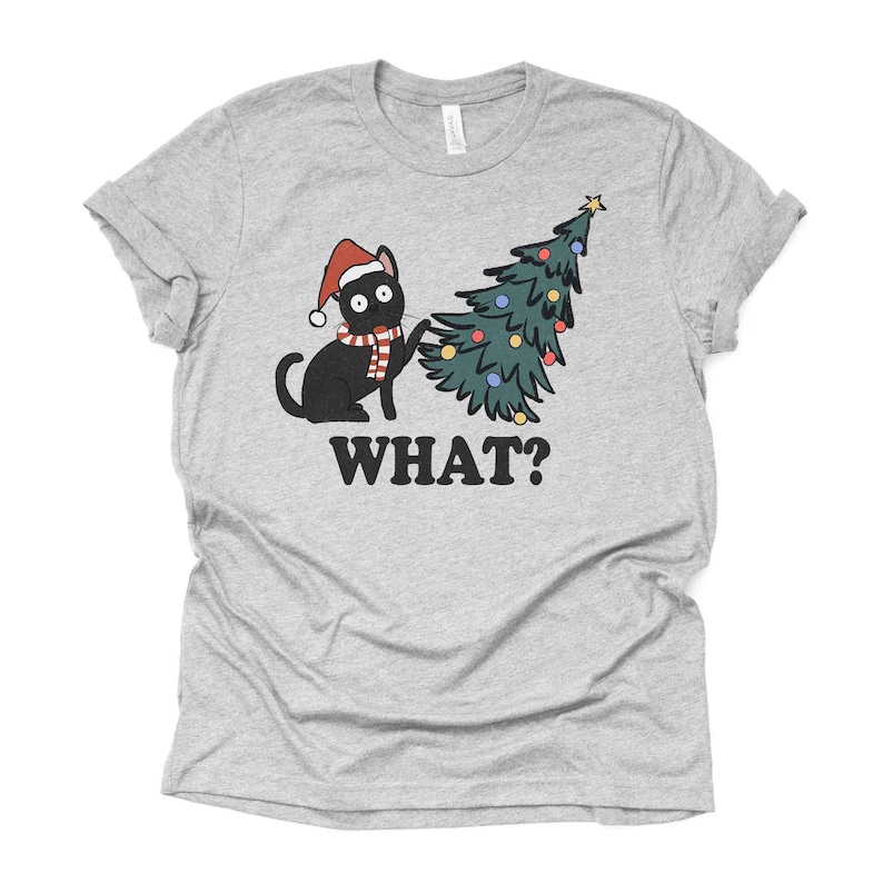 May include: A gray t-shirt with a black cat wearing a Santa hat and scarf, looking surprised, next to a green Christmas tree with colorful ornaments. The text "WHAT?" is printed below the image.