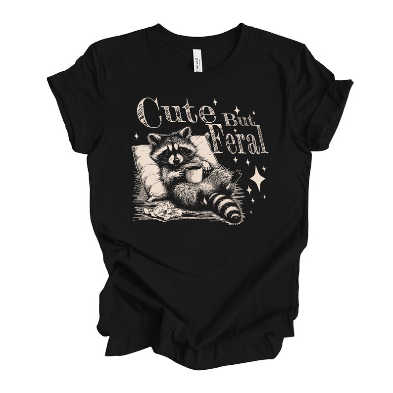 May include: Black t-shirt with a graphic of a raccoon holding a cup of coffee and the text "Cute But Feral".