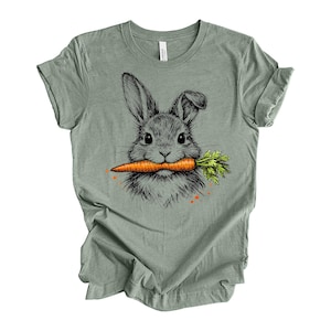 May include: A sage green t-shirt featuring a detailed illustration of a rabbit holding a carrot in its mouth. The rabbit is depicted in black and gray, with the carrot in orange and green. The shirt is made of a soft material.