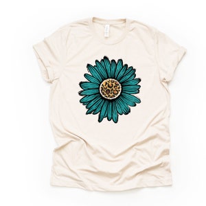 K&ouml;nnte beinhalten: Ein beiges T-Shirt mit einer t&uuml;rkisblauen Sonnenblume mit einem Leopardenmuster in der Mitte.