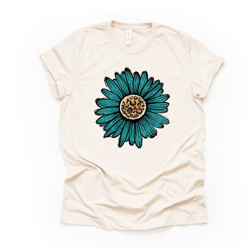 K&ouml;nnte beinhalten: Ein beiges T-Shirt mit einer t&uuml;rkisblauen Sonnenblume mit einem Leopardenmuster in der Mitte.