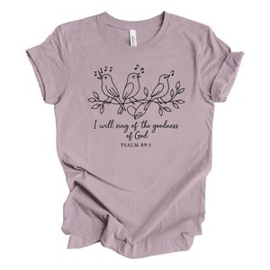May include: A mauve t-shirt featuring a black line drawing of three birds perched on a leafy branch, with musical notes above them. The text "I will sing of the goodness of God" and "Psalm 89:1" are below the birds.