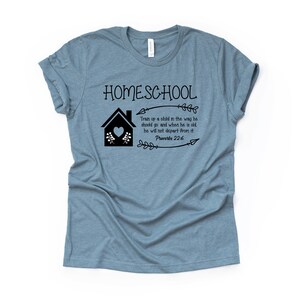 Puede incluir: Una camiseta azul claro con el texto "HOMESCHOOL" y una casa negra con un coraz&oacute;n. El texto "Train up a child in the way he should go and when he is old, he will not depart from it. Proverbs 22:6" tambi&eacute;n est&aacute; impreso en la camiseta.