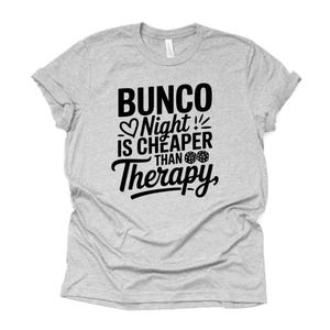 K&ouml;nnte beinhalten: Ein meliertes graues T-Shirt mit dem schwarzen Text "Bunco Night is Cheaper Than Therapy". Das Design umfasst ein Herz, W&uuml;rfel und Ausrufezeichen. Das T-Shirt ist aus weichem Material und hat kurze &Auml;rmel.