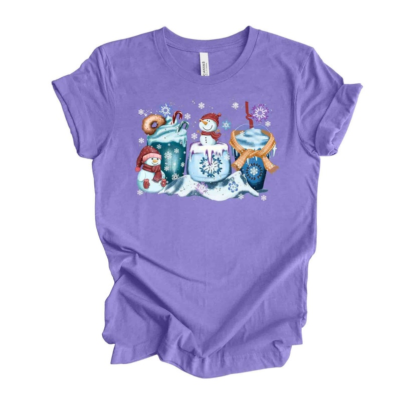 May include: Purple t-shirt with a winter-themed design featuring three cups of hot chocolate with whipped cream, a snowman, and snowflakes.