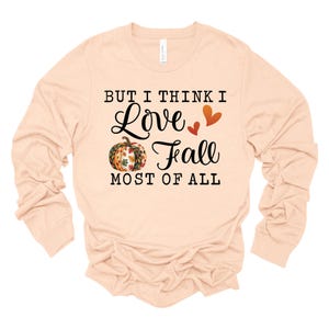 May include: A long-sleeved, peach-colored shirt with the text "BUT I THINK I Love Fall MOST OF ALL". The design includes a pumpkin with fall leaves and two hearts. A casual top for autumn.