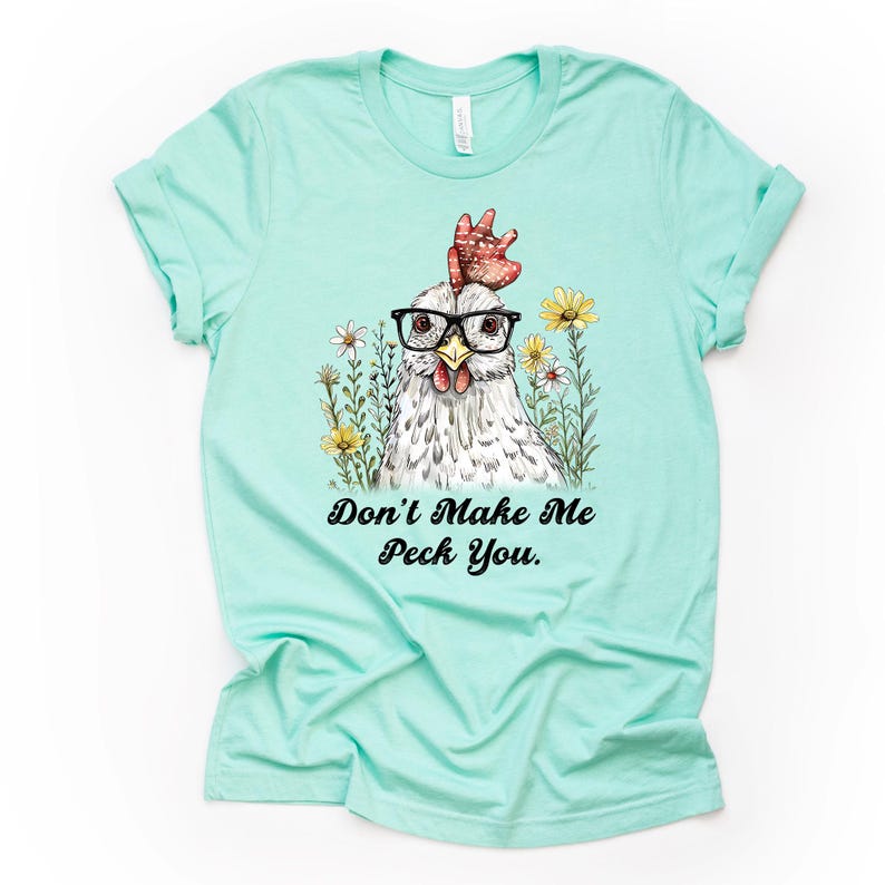 May include: A light teal t-shirt featuring a whimsical illustration of a chicken wearing glasses and a red comb, surrounded by flowers. The text "Don't Make Me Peck You" is below the image.