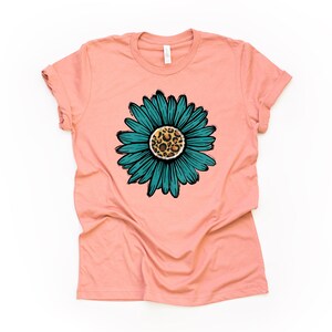 K&ouml;nnte beinhalten: Ein pfirsichfarbenes T-Shirt mit einer blaugr&uuml;nen  Margerite mit einem Leopardenmuster in der Mitte.