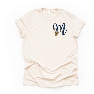 May include: A beige t-shirt with a navy blue letter "M" with gold accents and a sunflower design.