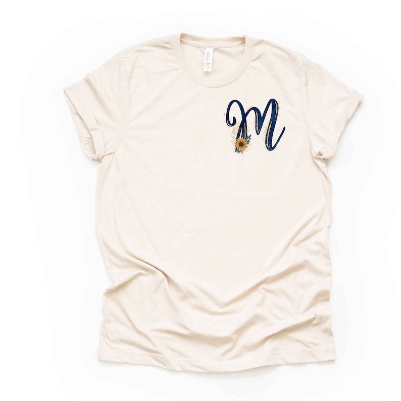 May include: A beige t-shirt with a navy blue letter "M" with gold accents and a sunflower design.