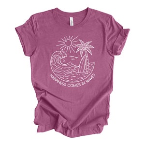 May include: A mauve t-shirt with a white graphic of a wave, palm tree, and sun. The text "Happiness comes in waves" is printed below the graphic.