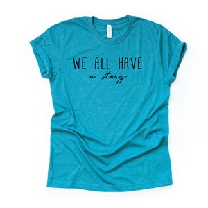 May include: A teal blue t-shirt with the text "WE ALL HAVE a story" printed in black.