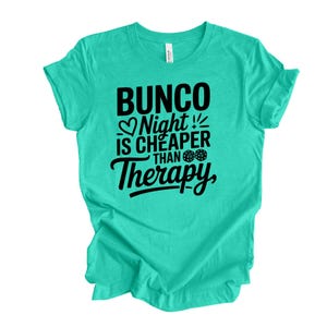 K&ouml;nnte beinhalten: Ein t&uuml;rkisfarbenes T-Shirt mit dem Text "Bunco Night is Cheaper Than Therapy" in schwarzer Schrift. Das Design beinhaltet ein Herz, W&uuml;rfel und Ausrufezeichen. Das Shirt ist aus einem weichen Material.
