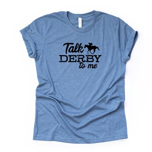 Peut inclure: Un t-shirt bleu clair avec le texte "Talk Derby to me" en lettres noires. Une silhouette noire d'un jockey &agrave; cheval est au-dessus du mot "Derby".