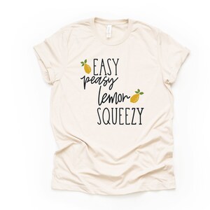 Puede incluir: Una camiseta beige con el texto "EASY peasy lemon SQUEEZY" en letra cursiva negra. Dos limones amarillos con hojas verdes est&aacute;n incluidos en el dise&ntilde;o.